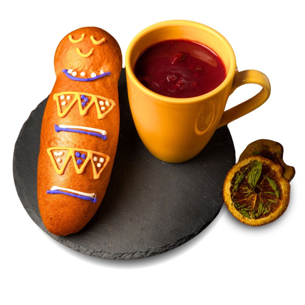 Guagua de pan decorada junto a una taza amarilla con bebida morada, sobre una base oscura.
